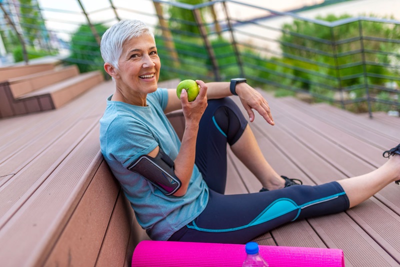 older woman holding an apple sitting next to a yoga mat