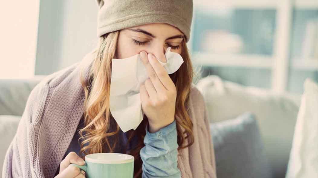 A sick woman holding a mug blows her nose with a tissue.