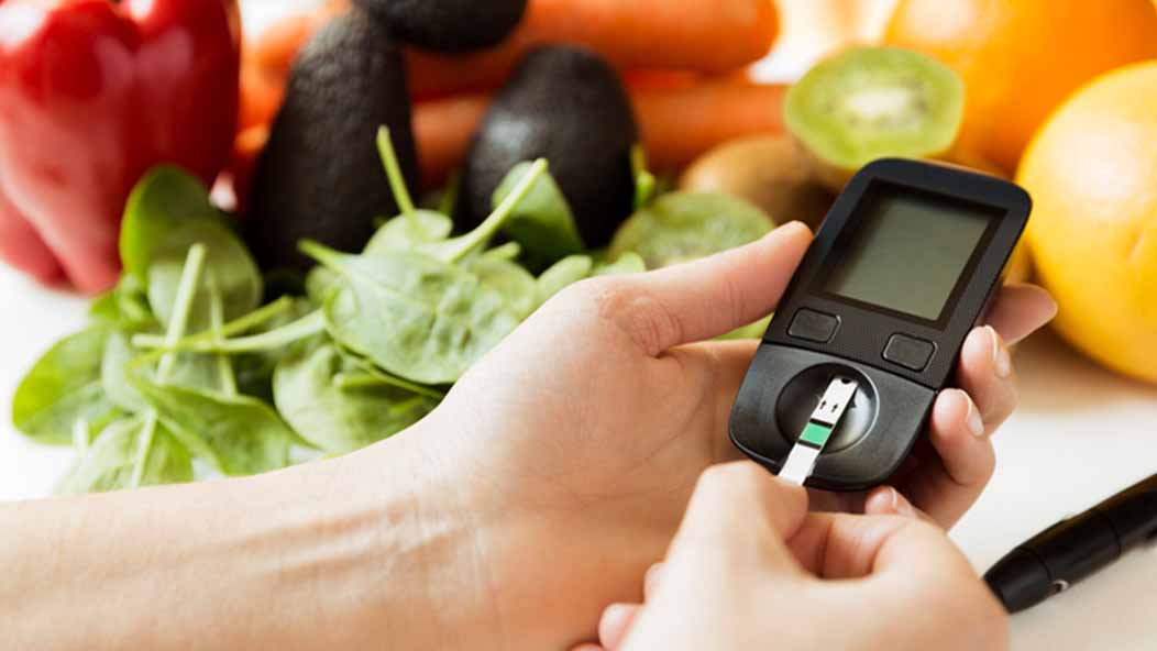 Close-up of a person inserting a test strip into a blood glucose meter with assorted fresh, colorful vegetables in the background.