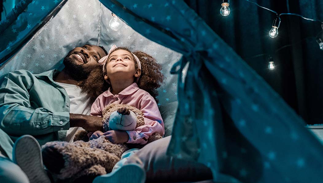 A father and his daughter, in pajamas and holding her teddy bear, look up smiling as they sit close together in a tent made of blankets.