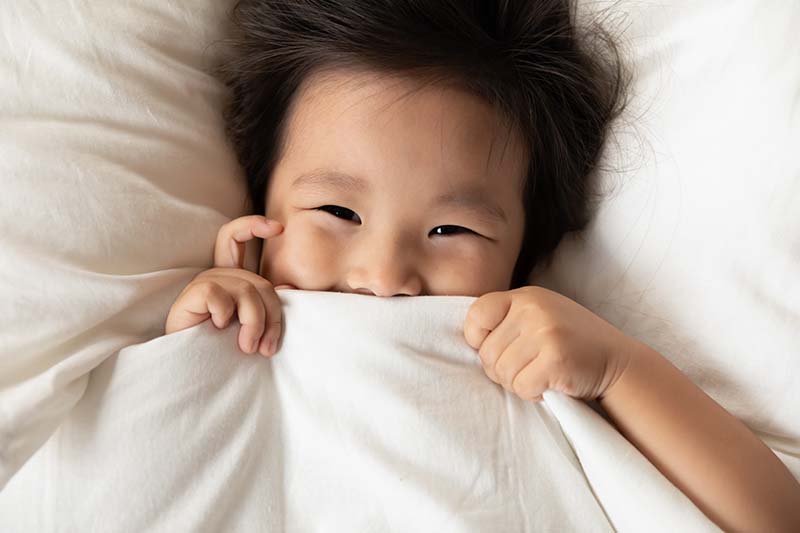 A young child pulls sheets up over the bottom half of its face while smiling.