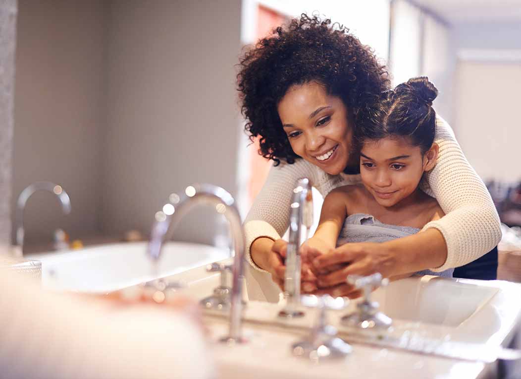 A mother standing behind her young daughter reaches her arms around while helping the child wash her hands in the sink.