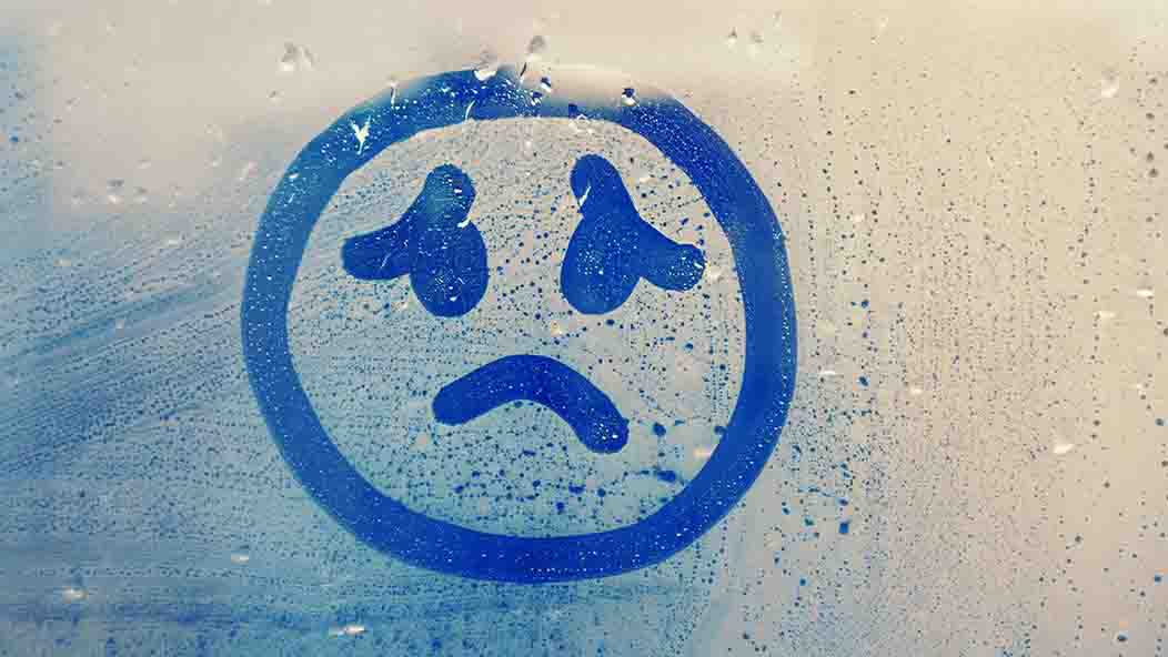 A simple picture of a sad face, presumably drawn with a finger, is drawn on a window lined with condensation.
