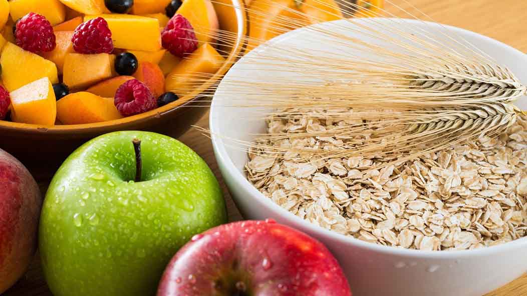 High-fiber foods such as red and green apples, pineapple chunks in a bowl with berries, and a bowl of oats are displayed on a countertop.