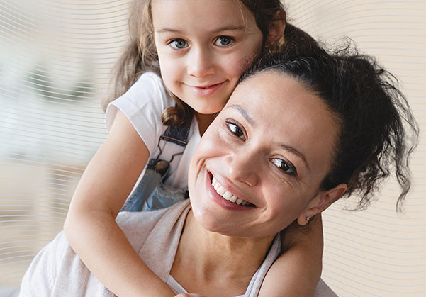 A young girl wraps her arms around her mom's shoulders as they both smile.